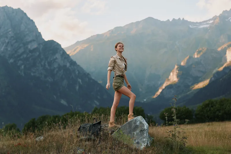 Confident woman enjoying the outdoors in a mountain meadow, casual outfit, sunlight on her face, posing with a rock, expansive valley behind, lifestyle moment in nature