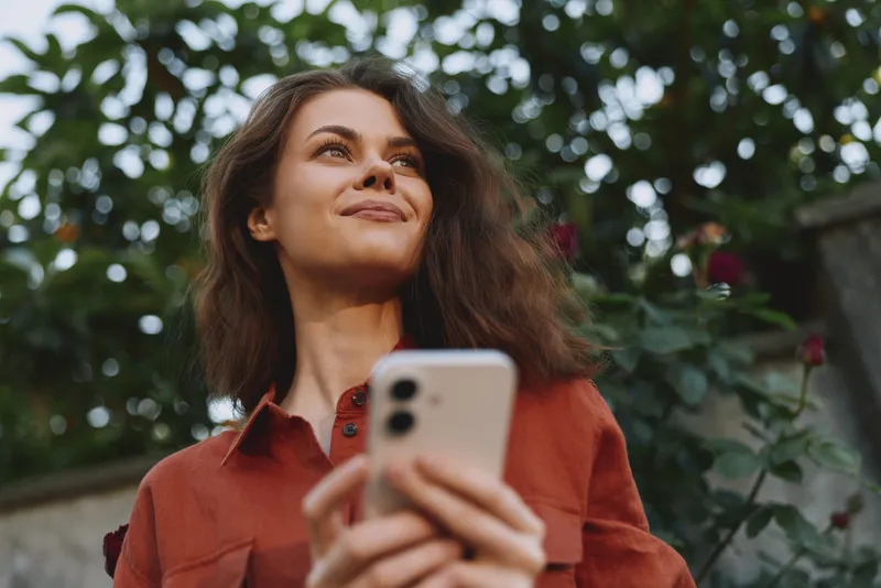 Young woman in a rust shirt holds a smartphone outdoors, smiling softly as green foliage frames her, conveying everyday connectivity and relaxed confidence