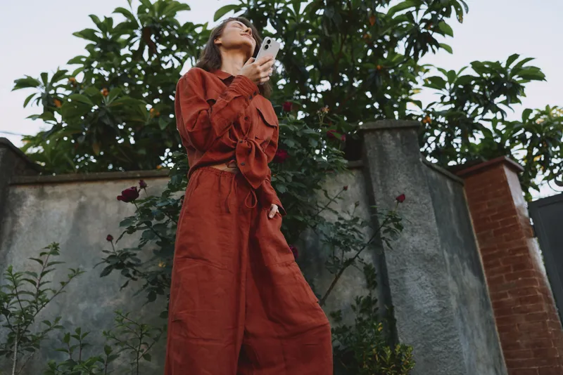 Cheerful woman outdoors in a rust red dress, looking up with a phone in hand, sunlit breeze, relaxed lifestyle moment, natural colors, lush greenery and urban garden backdrop