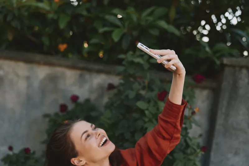 Joyful woman outdoors, taking a selfie with a smartphone, smiling at the camera in casual attire, warm sunlight, lush greenery, relaxed mood, vibrant lifestyle moment