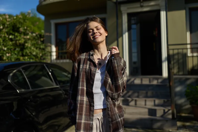 Young woman outdoors wears a plaid shirt over a white top, standing in bright sunlight with a relaxed smile and carefree mood, her hair moving in a light breeze