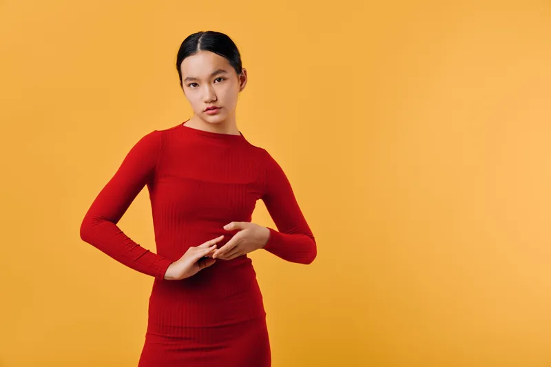 Young woman in red dress poses with crossed hands against orange background for fashion, style, beauty, and confident model portrait in studio with minimalistic design and vibrant color.