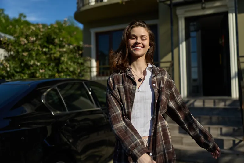 Smiling woman in a plaid shirt walks outdoors near a house, showcasing a casual lifestyle and relaxed confidence. Natural light highlights her cheerful expression. Bright daylight adds warmth
