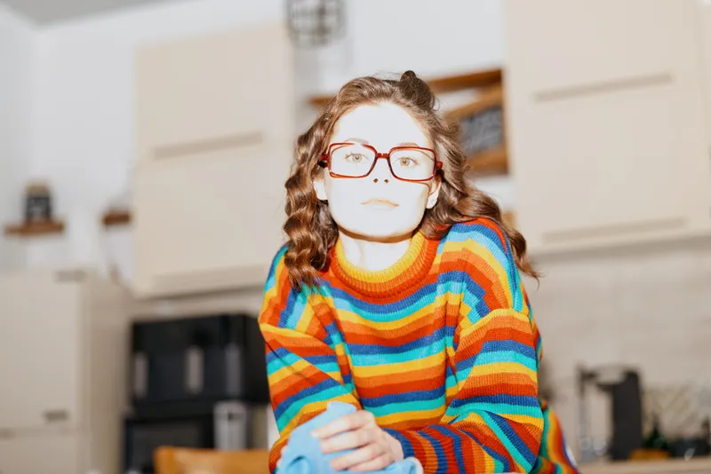 Woman in a colorful striped sweater and red glasses sits indoors in a bright home kitchen, presenting a relaxed, thoughtful lifestyle moment with warm daylight and cozy mood.