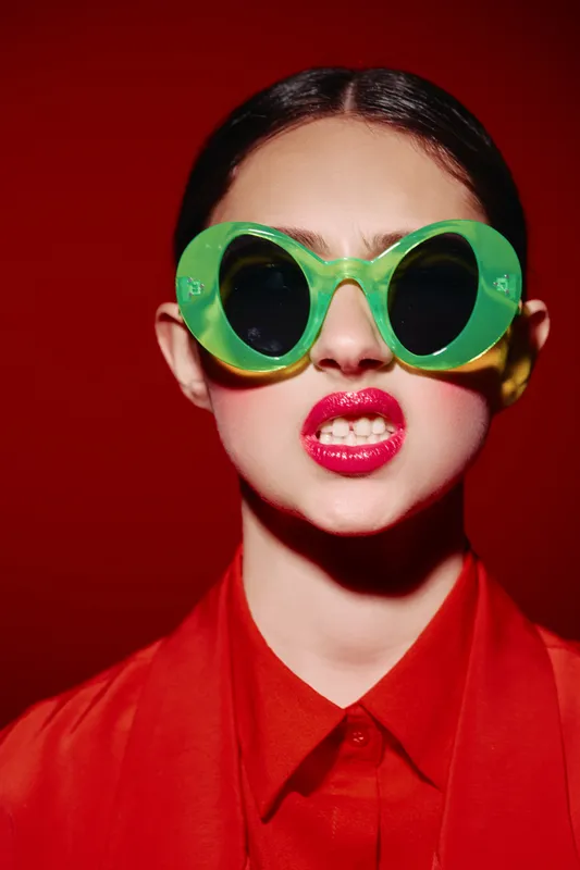 Young woman wearing oversized green sunglasses with red lipstick, expressive face and bold fashion styling on vivid red background. Concept for beauty creative fashion statement.