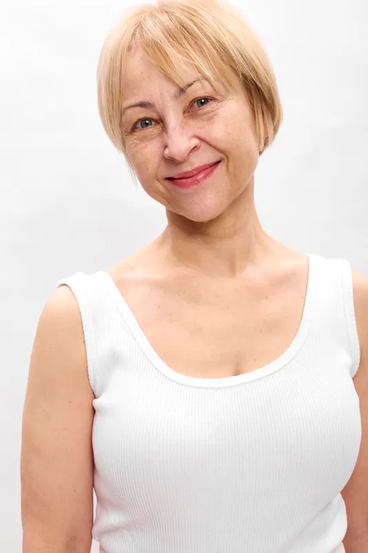 Elderly woman portrait, blonde short hair, gentle smile, white sleeveless top, relaxed posture, clean white background, soft studio light, neutral expression