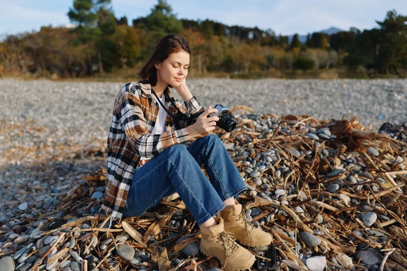 Woman photographer sits on a rocky beach during autumn, holding a camera and focusing on composition amid pebbles and driftwood, casual plaid jacket and denim jeans for an outdoor shoot.