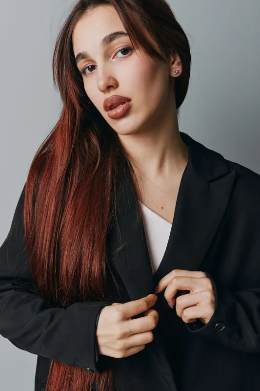 Woman with long hair wearing black blazer and white top posing against gray background. Female model with natural makeup and serious expression looking at camera indoors.