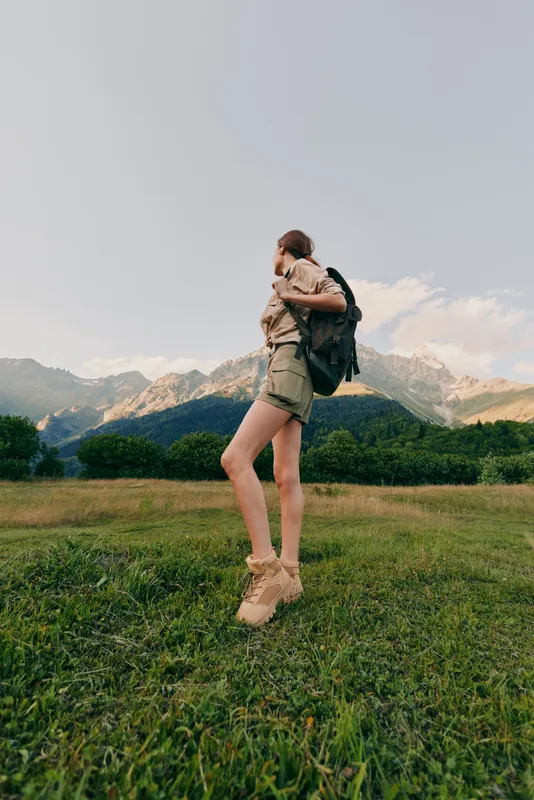 Young woman hiking in a grassy field with mountains in the background, wearing a backpack and travel outfit, smiling as she explores the outdoors on a sunny day