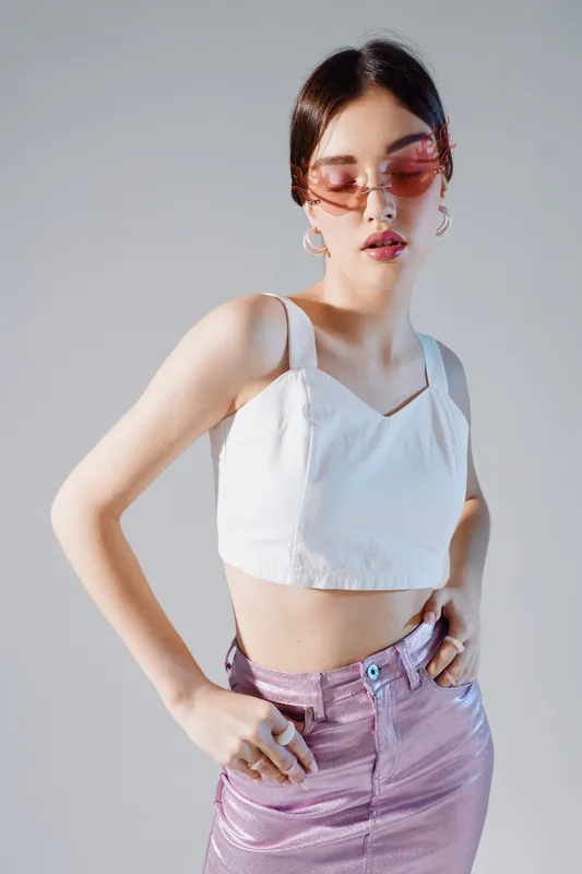 Young woman in a white crop top and shiny pink skirt poses confidently wearing pink sunglasses. Studio shot on a neutral background. Concept of woman beauty fashion and style.