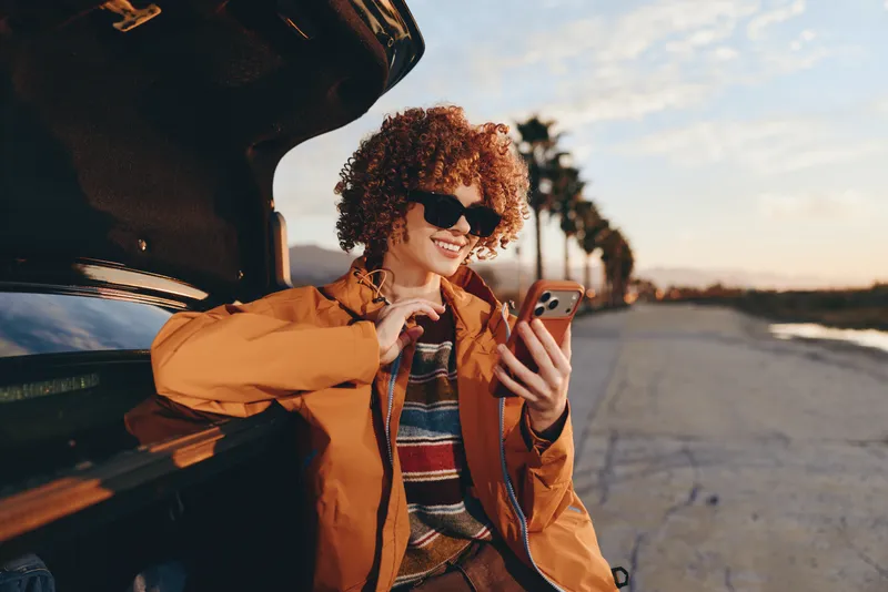 Woman lifestyle outdoors by car at sunset, smiling with curly hair, holding smartphone near open trunk, warm golden light, casual orange jacket, relaxed street moment, natural colors
