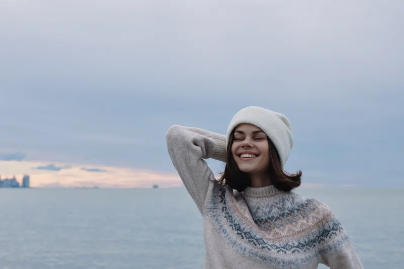 Woman in a knit sweater and beanie smiles by the ocean on a cool day, wind in her hair, calm seascape behind, relaxed outdoor portrait showcasing winter style and contentment