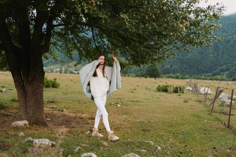 Young woman in a white knit sweater and pants poses beneath a leafy tree in a rural field, capturing a relaxed, carefree lifestyle moment.