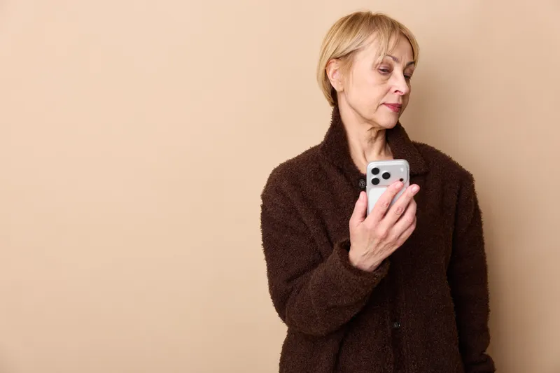 Senior woman holding smartphone in studio, beige background, she checks the screen using technology for communication and social media, calm mood, soft studio light