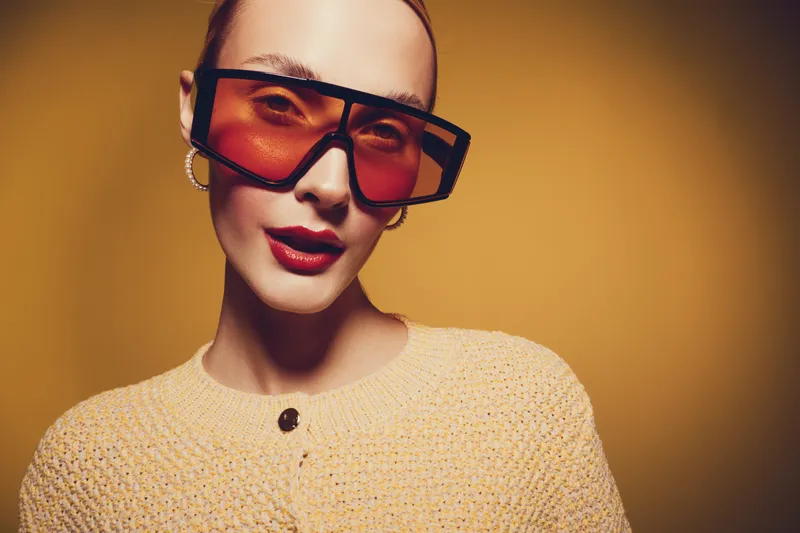 Stylish woman with oversized orange sunglasses and red lipstick poses confidently against a warm yellow background. Fashion concept with bold accessories and makeup.