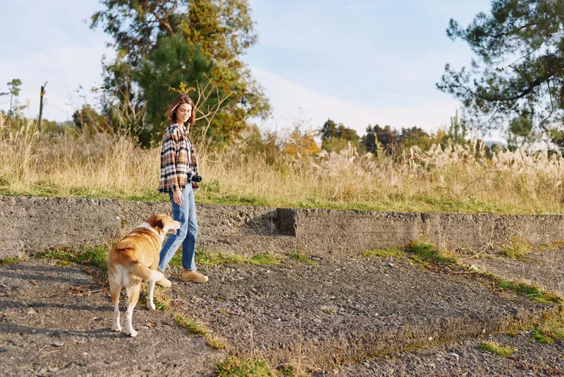 Woman walking a dog along a gravel path in a sunny park, casual attire, relaxed outdoor activity, nature in the background, everyday companionship