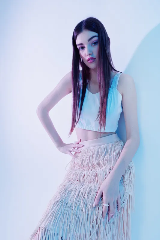 Young woman beauty fashion model poses with long hair in a white top and textured skirt in soft studio lighting on a light background.