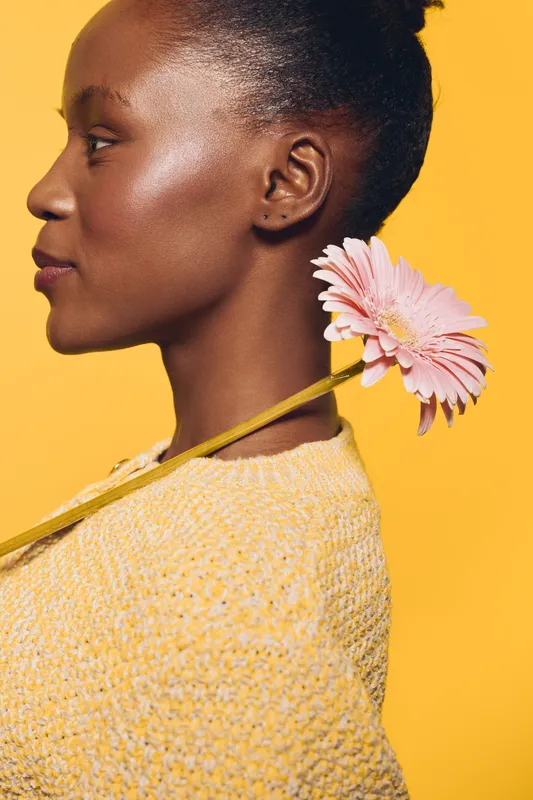 Profile of a young black African woman with dark skin wearing a yellow sweater, holding a pink gerbera flower behind her neck on a bright yellow background. Concept of beauty and nature.