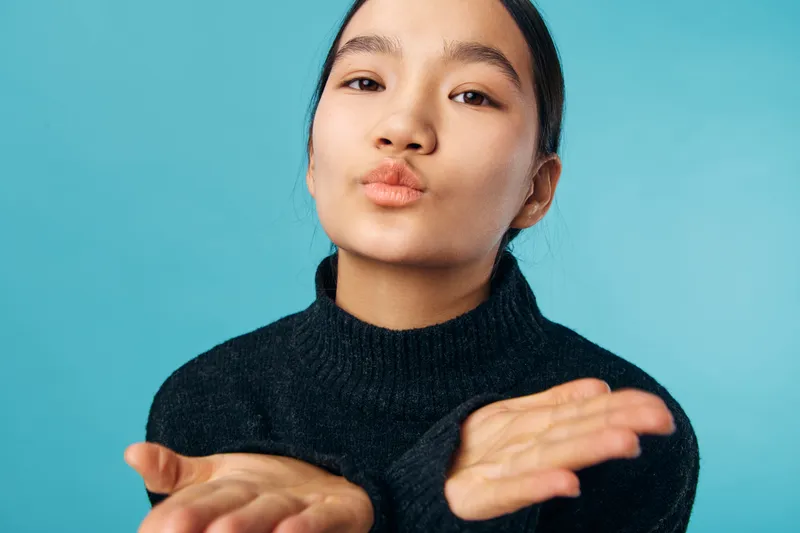 Young woman in black sweater blowing kiss with hands extended forward against blue background. Portrait of beauty, affection, gesture, and friendly emotion expressing love or hello.