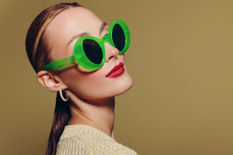 Stylish woman with slicked back hair wears large green sunglasses and red lipstick. She poses confidently against a plain beige background. Concept of fashion and modern style.