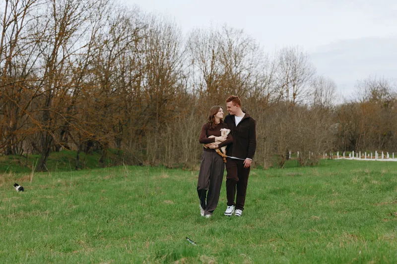 Couple outdoors in a park on a sunny autumn day, standing on grass among trees, smiling and relaxed, holding a pet, candid lifestyle moment in natural light