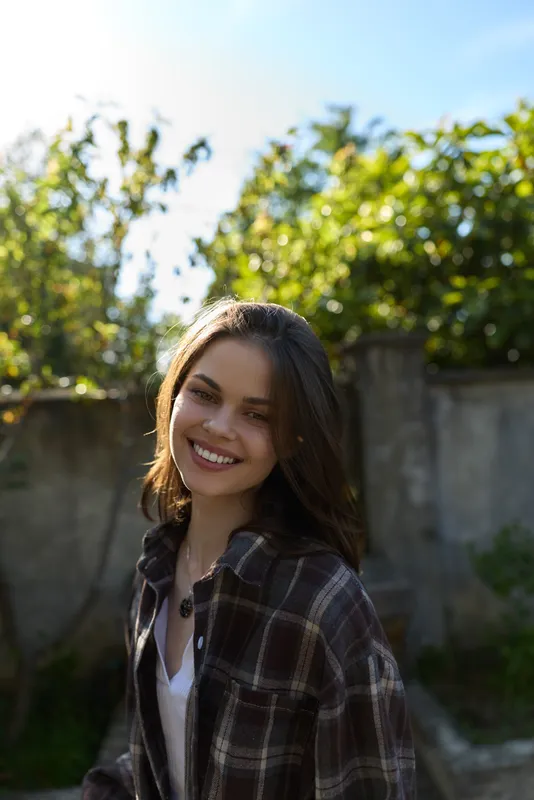 Young woman with brunette hair smiles warmly in a sunny outdoor setting, wearing a plaid shirt over a white tee, relaxed and confident, with soft natural light and greenery behind