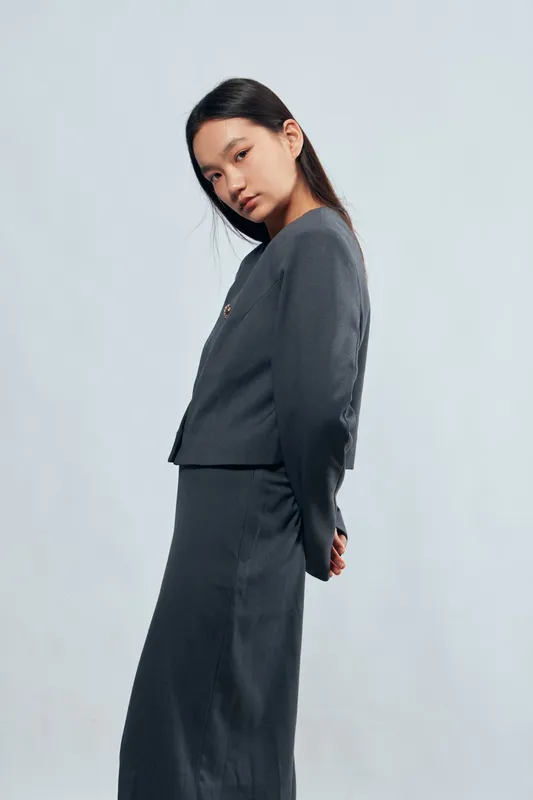 Young woman in dark gray long sleeve top and skirt with hands behind back standing in studio with plain background, looking at camera with serious expression and straight hair.