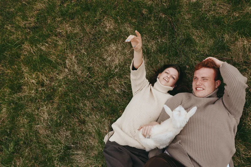 A couple lying on grass outdoors, smiling with a white goat kid between them, capturing a playful lifestyle moment of affection and rural joy, warm daylight enhancing connection and togetherness.