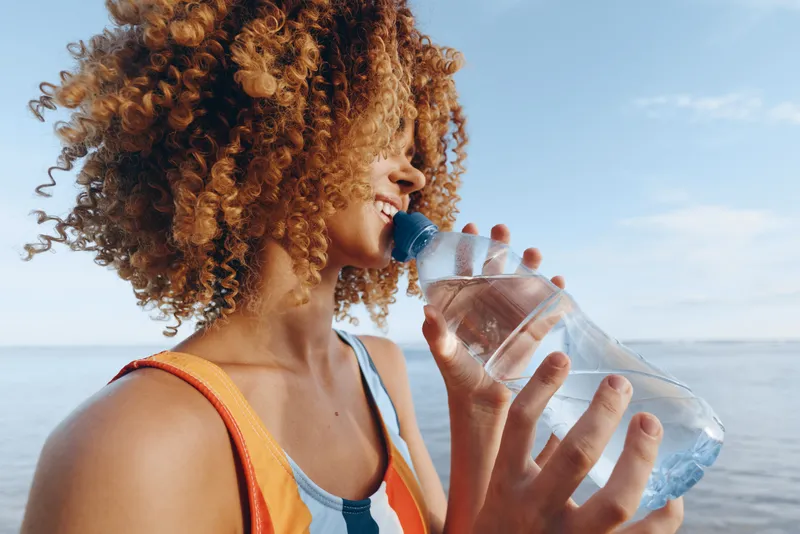 Woman with curly hair drinks water from a transparent bottle, near the ocean in summer, outdoors, focused on hydration and refreshment, healthy lifestyle for thirst relief and wellbeing
