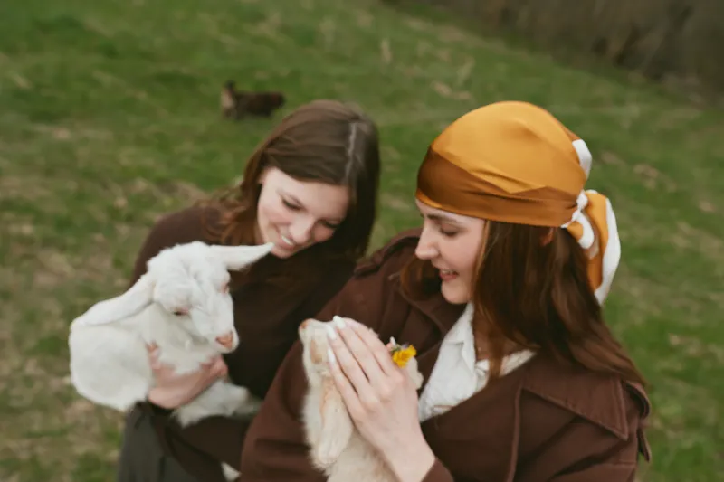 Cheerful women outdoors, two friends share a gentle moment with a small goat, kid, soft light, warm colors, grassy field, relaxed interaction, candid lifestyle scene, affectionate dozen smiles