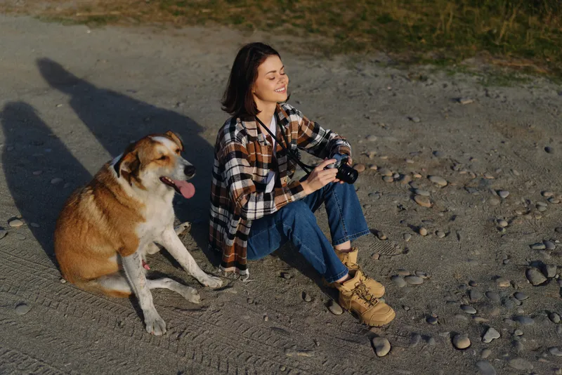 Woman sits on a rocky path with a dog beside her, holding a camera. Casual plaid shirt, boots and warm sunlight create a friendly outdoor photography portrait