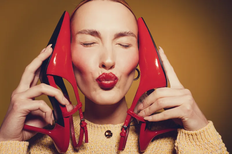 Young woman with closed eyes holds red high heels near face and makes kiss gesture. Studio portrait on yellow background. Concept of fashion, beauty, and style accessories.