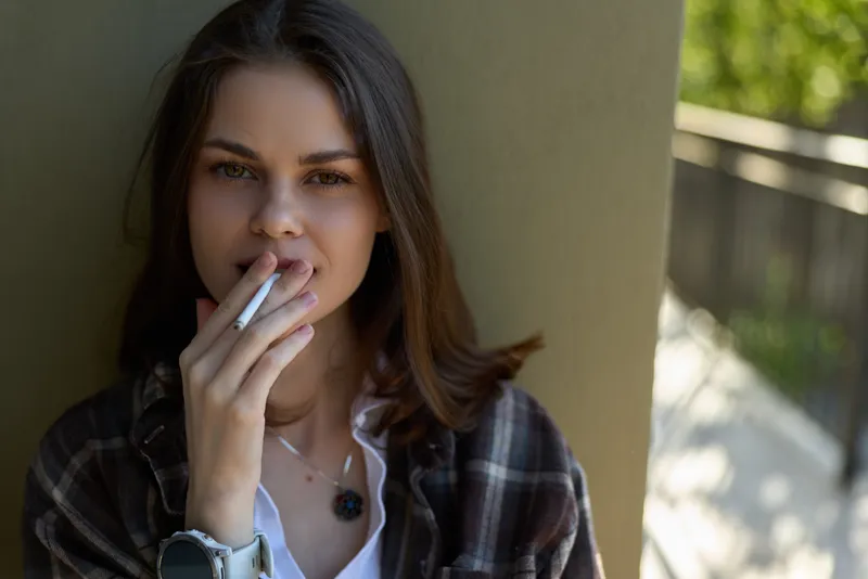 Smiling woman outdoors, smoking a cigarette, wearing a plaid shirt, casual lifestyle scene, natural daylight, relaxed mood, candid portrait in a sunny courtyard