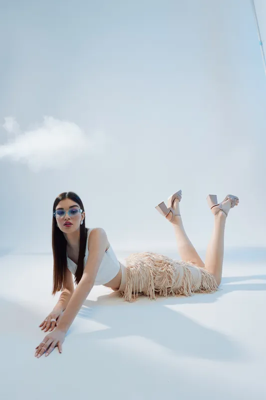 Fashionable woman with long dark hair wearing sunglasses and a fringed skirt poses lying on the floor against a bright sky background. Concept of beauty and fashion.