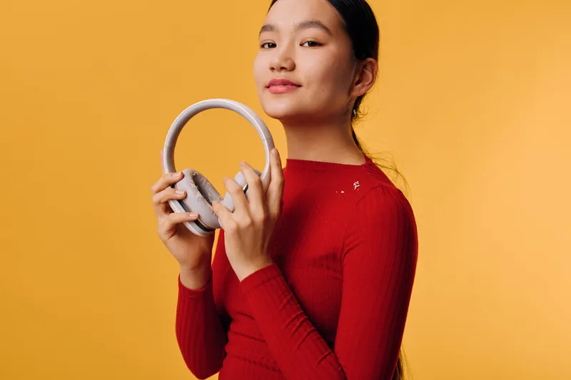 Young woman holding wireless headphones and looking at camera against orange studio background. Portrait of confident girl with red sweater and casual hairstyle in modern tech accessory concept.