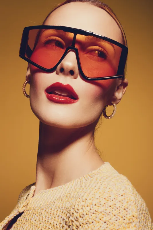Stylish woman with red lips and oversized orange sunglasses poses against yellow background. Fashion portrait with hoop earrings and knitted top. Concept of modern style and beauty.