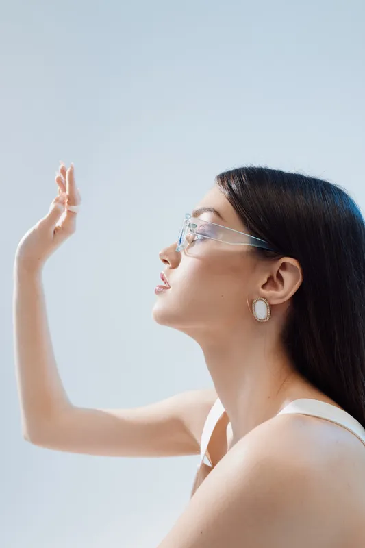 Profile of a beautiful woman with long dark hair wearing transparent glasses and white earrings, posing with hand raised against a light background. Concept of beauty fashion.