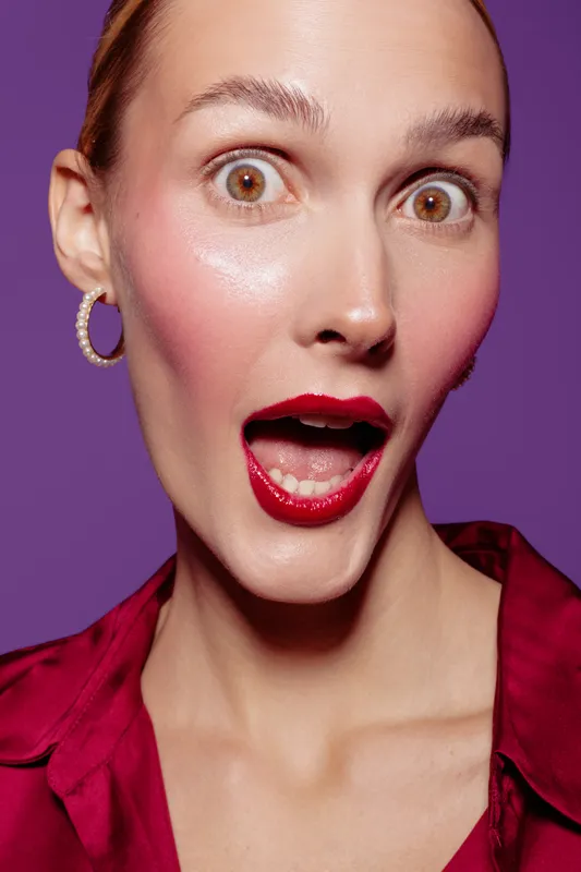Surprised woman with wide eyes and open mouth wears red lipstick and earrings. Close-up portrait on purple background. Concept of shock, emotion, and beauty expression.
