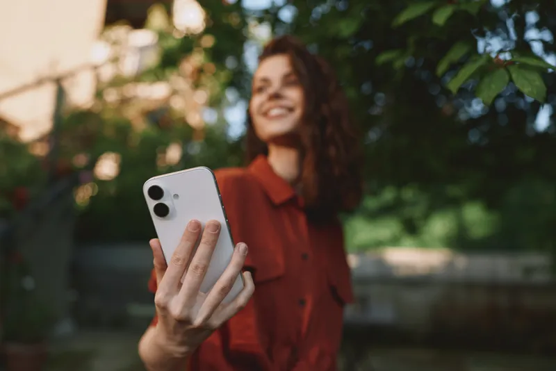 A young woman in a red shirt takes a selfie outdoors, smiling into the camera as she enjoys a casual, sunlit moment in a leafy garden, embodying everyday lifestyle