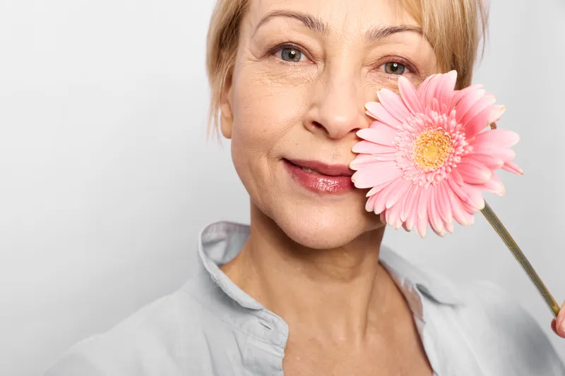 woman portrait gerbera flower, beauty skincare smile in studio closeup, senior woman gently holds pink blossom by her lips, soft light, white background, calm elegance