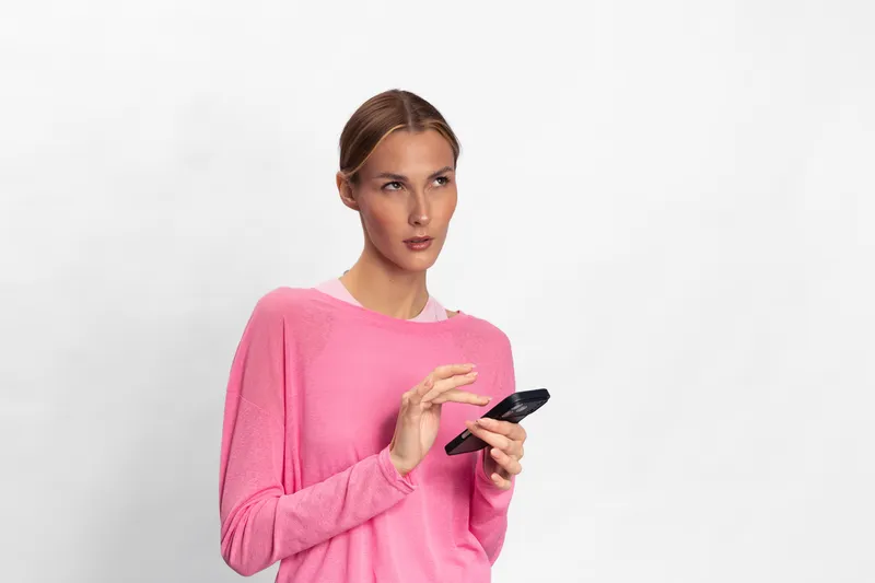Young woman in pink sweater looks away while holding smartphone in hand. Studio portrait with white background. Concept of communication, technology, and mobile device use.