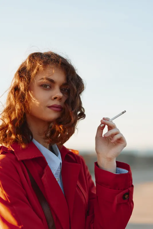 Young woman with curly red hair wearing a red coat holds a cigarette outdoors while looking confidently at the camera during golden hour.