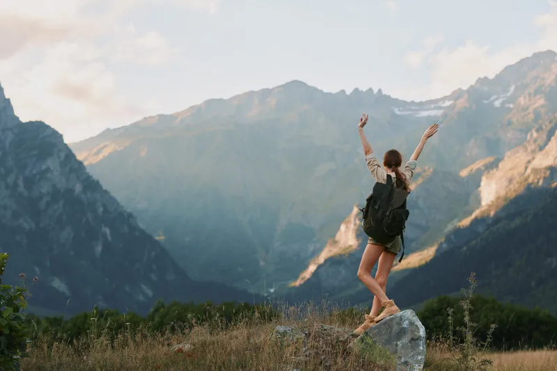Young woman with a backpack stands on a rock in a dramatic mountain landscape, arms raised in triumph, capturing a carefree outdoor adventure moment today.