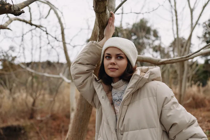 Woman in a beige winter coat and knit hat stands among leafless trees, posing calmly in an outdoor forest setting, capturing a cozy, minimalist vibe and a serene moment of seasonal solitude.