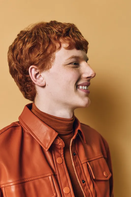 Young man with red hair wearing brown leather jacket and turtleneck smiling in profile against beige background. Curly hairstyle and casual fashion with happy expression and natural light.