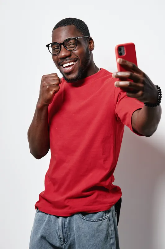 African American man with dark skin wearing glasses and red t shirt taking selfie with smartphone while smiling and making fist gesture in studio with white background.
