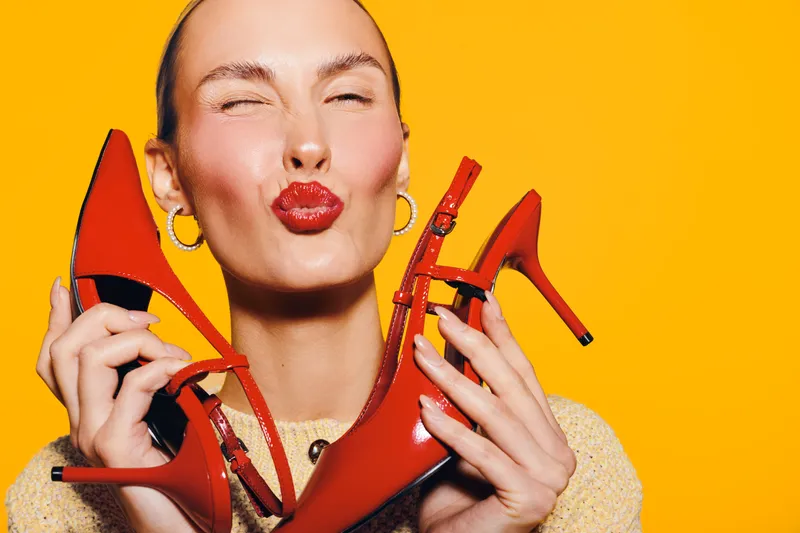 Happy woman with red lipstick holds red high heel shoes close to her face, eyes closed and lips puckered. Bright orange background creates vibrant and playful fashion concept.