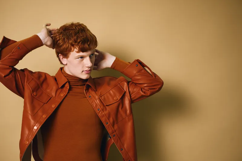 Young man with red curly hair wearing brown leather jacket and turtleneck posing with hands behind head in studio with beige background for fashion and style.