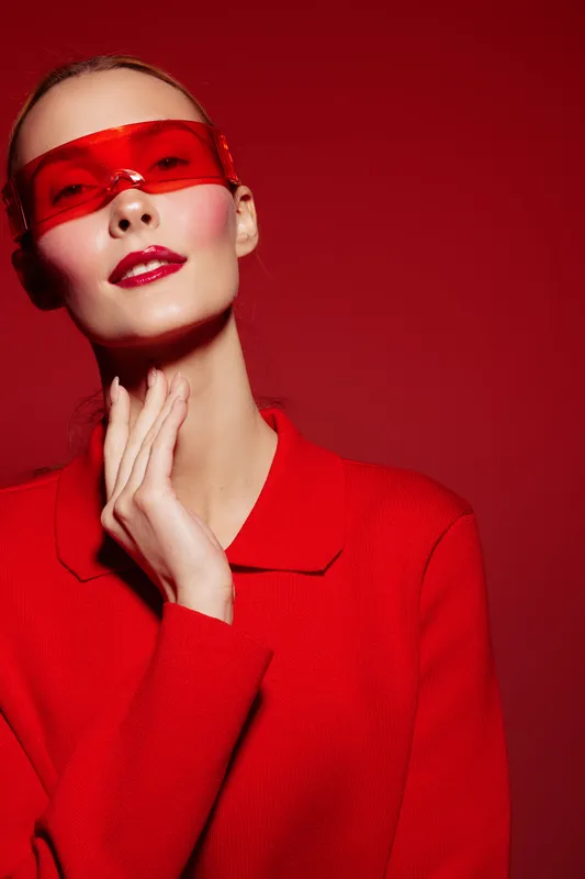 Stylish woman in red sweater and red translucent glasses poses against red background. Fashion portrait with bold colors and modern accessories, concept of style and confidence.