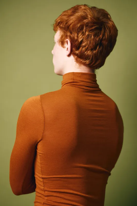 Young man with red hair wearing brown turtleneck sweater standing turned back against olive green background with arms crossed. Isolated studio portrait with soft lighting and casual style.